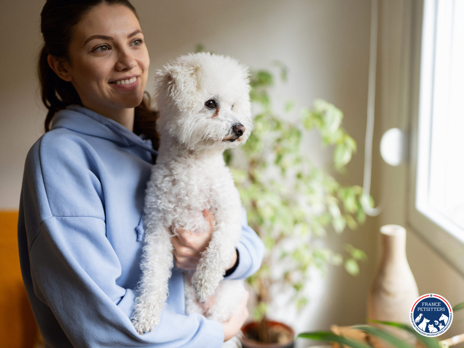 Une petsitter tient un bichon dans ses bras devant une fenêtre